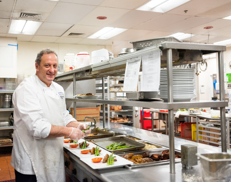 Chef Anthony Napolitano smiling while plating asparagus and tomatoes in a professional kitchen at Edgehill, with trays of prepared dishes and stainless steel counters in the background.