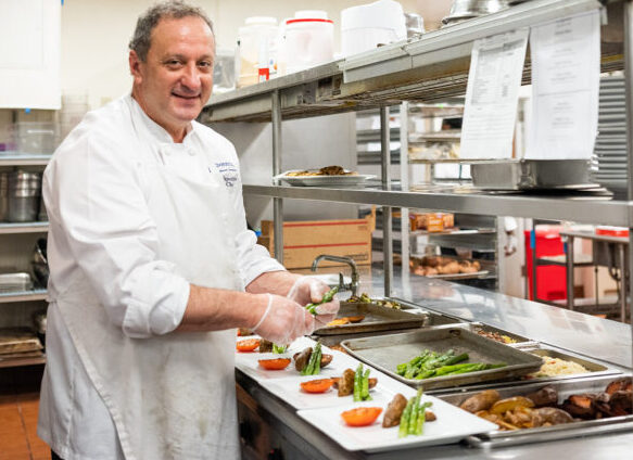 Chef Anthony Napolitano smiling while plating asparagus and tomatoes in a professional kitchen at Edgehill, with trays of prepared dishes and stainless steel counters in the background.