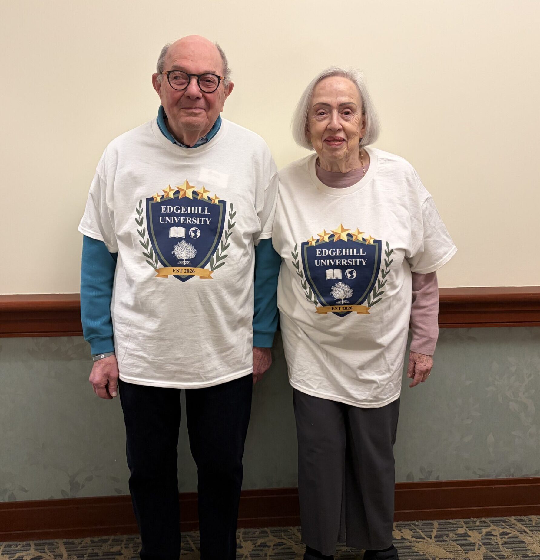 Ron and Susan Ebert standing side by side indoors, smiling and wearing Edgehill University T-shirts that celebrate the resident-led lifelong learning program.