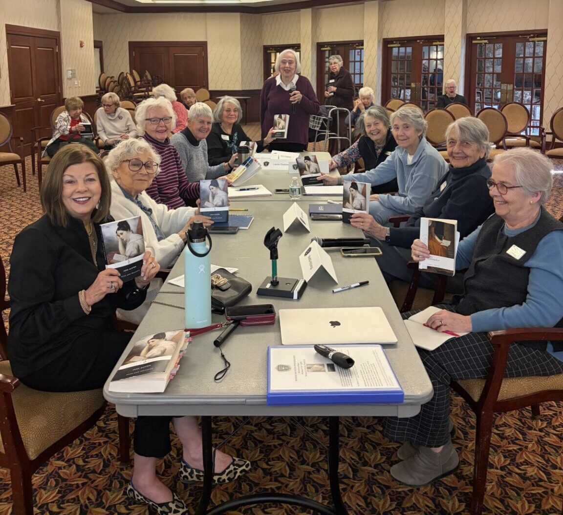 A group of Edgehill residents gathered around a table during a class, smiling and holding books as they participate in a lively learning session.