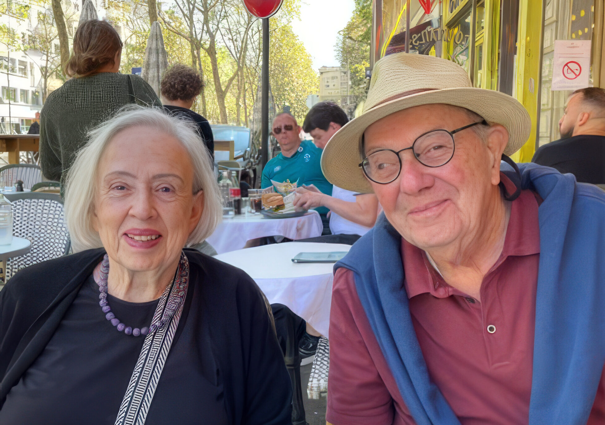 An older couple smiling while seated at an outdoor café table, enjoying time together on a sunny day with trees and other diners in the background.