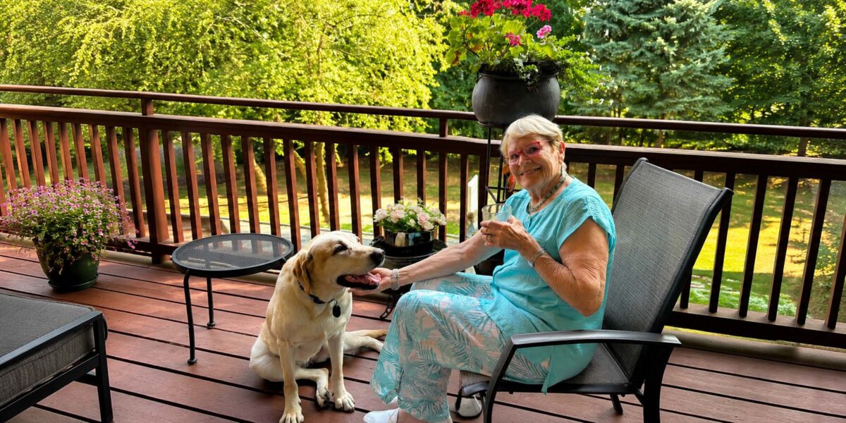 A smiling older woman relaxes on a sunny outdoor deck with her light-colored Labrador retriever by her side. Surrounded by potted flowers and lush green trees, she sits in a patio chair holding a drink while petting the dog in a peaceful residential setting.