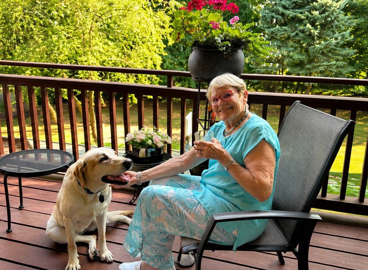 A smiling older woman relaxes on a sunny outdoor deck with her light-colored Labrador retriever by her side. Surrounded by potted flowers and lush green trees, she sits in a patio chair holding a drink while petting the dog in a peaceful residential setting.